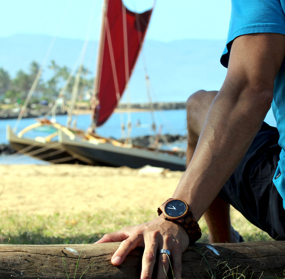 round koa wood watch on mans wrist sitting looking at Polynesian sailing canoe
