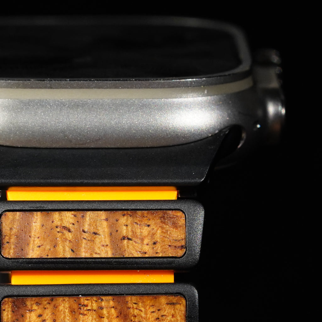 Close-up of a smartwatch with a dark metallic case and a wooden band featuring warm brown grain and bright orange accents, set against a black background.