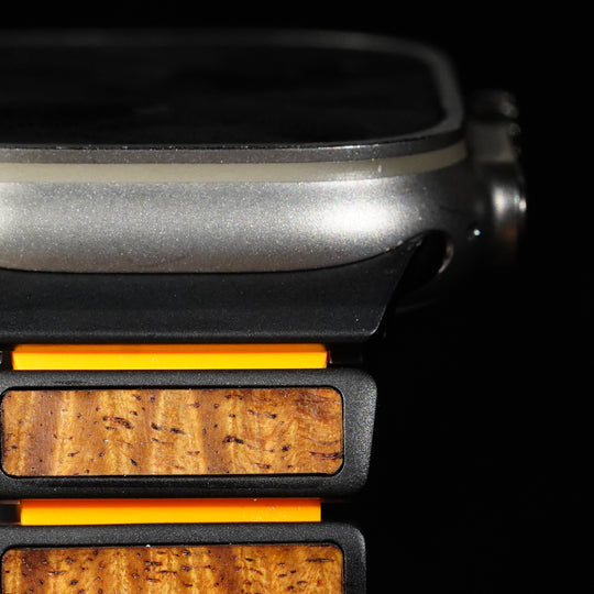 Close-up of a smartwatch with a dark metallic case and a wooden band featuring warm brown grain and bright orange accents, set against a black background.